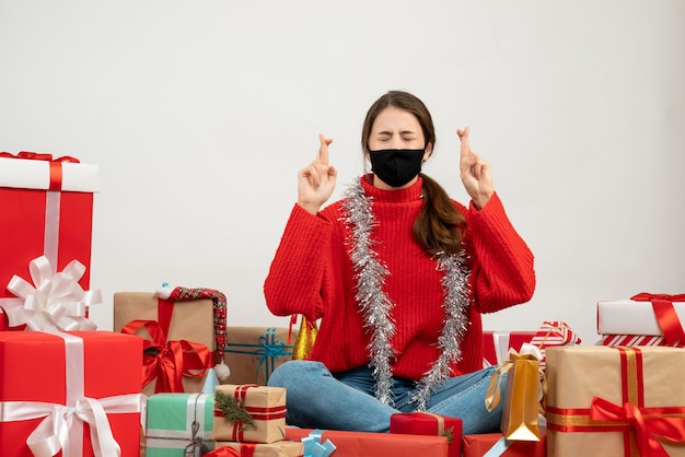 Niña con suéter rojo y máscara negra haciendo el signo de la buena suerte sentados alrededor de regalos en blanco