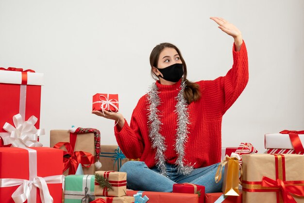 Niña con suéter rojo y máscara negra con regalo levantando su mano sentada alrededor de regalos en blanco
