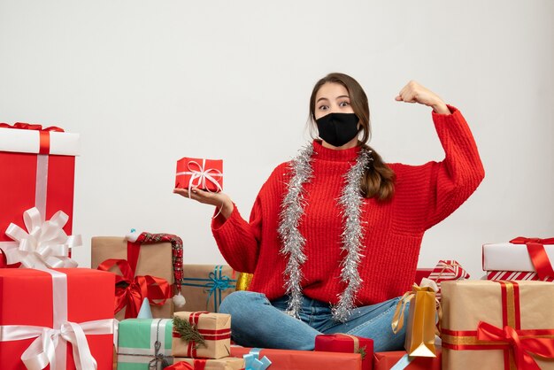 Niña con suéter rojo y máscara negra sosteniendo un regalo mostrando su fuerza sentada alrededor de regalos en blanco