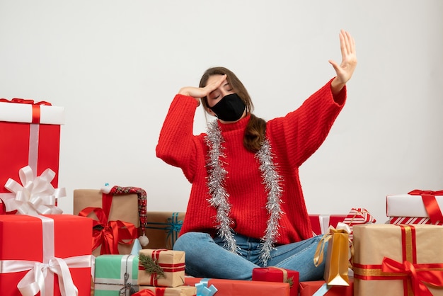 Niña con suéter rojo poniendo la mano en la frente sentado alrededor de regalos con máscara negra sobre blanco