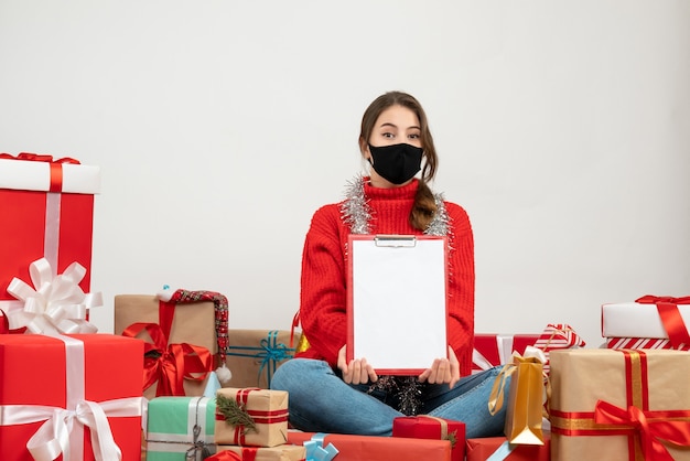 Niña con suéter rojo sosteniendo archivos sentados alrededor de regalos con máscara negra sobre blanco