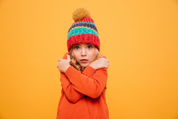 Niña en suéter y sombrero con frío y mirando a la cámara sobre naranja