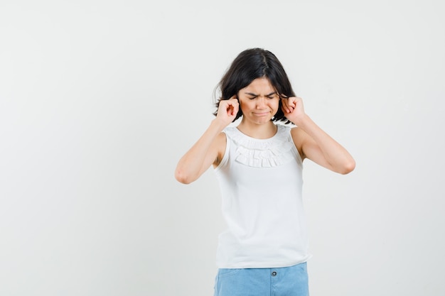 Niña tapando las orejas con los dedos en una blusa blanca, pantalones cortos y mirando molesto, vista frontal.