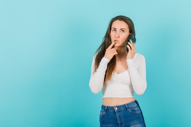 Niña con teléfono inteligente está pensando sosteniendo el dedo índice en los labios sobre fondo azul.