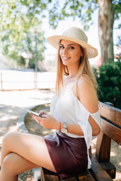 Niña con un teléfono inteligente y mensajes de texto sentado en un banco de un parque urbano