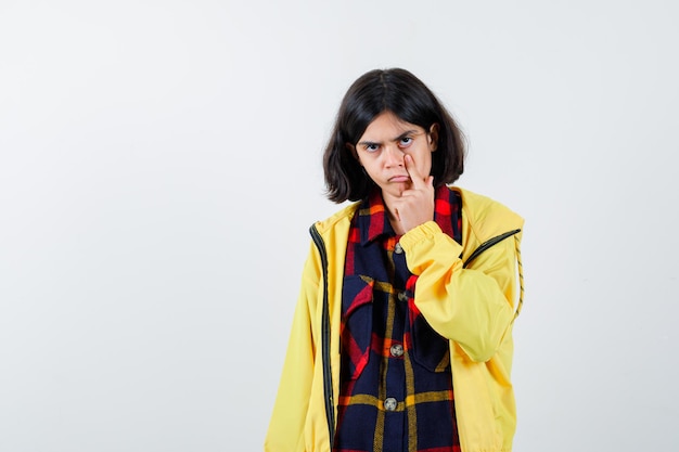 Niña tirando hacia abajo el párpado en camisa a cuadros, chaqueta y luciendo bonita, vista frontal.