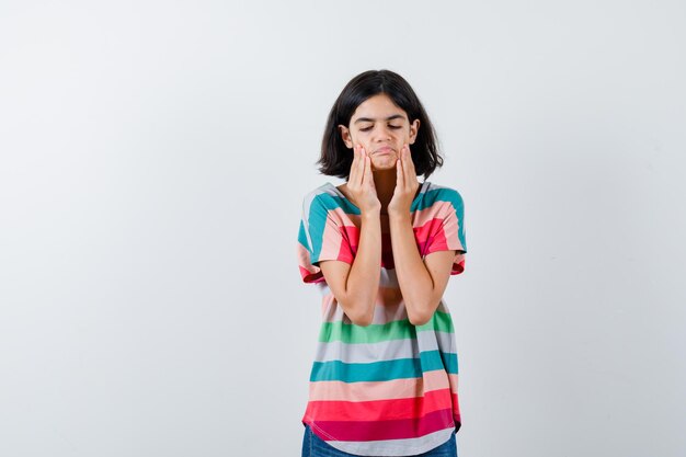 Niña tirando de la piel de las mejillas en camiseta y mirando molesto, vista frontal.