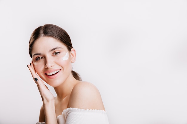 Niña toca la piel hidratada y sonríe. Retrato de modelo con crema en la cara en pared aislada.