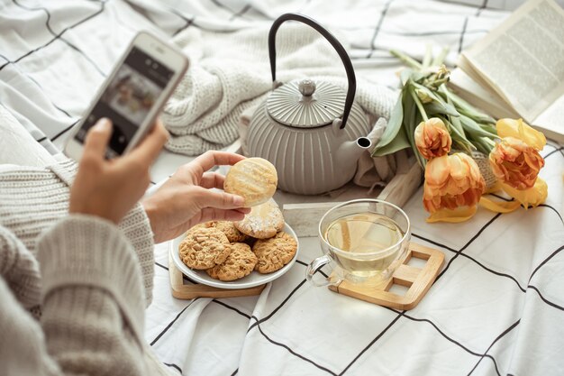 Una niña toma fotografías en el teléfono de una composición de primavera con té, galletas y tulipanes en la cama