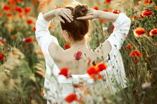 La niña toma las manos por la cabeza y se para con la espalda desnuda con una flor de tatuaje amapola, entre el campo de amapolas