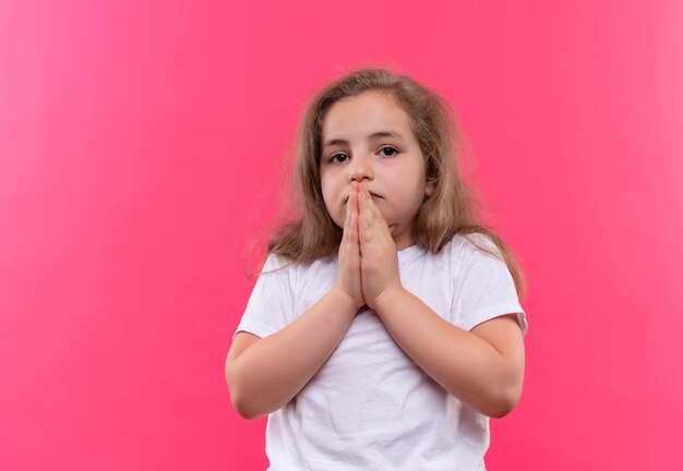 Niña triste de la escuela con camiseta blanca que muestra gesto de oración sobre fondo rosa aislado