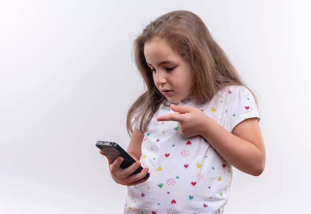 Niña triste de la escuela vistiendo camiseta blanca puntos en el teléfono sobre fondo blanco aislado