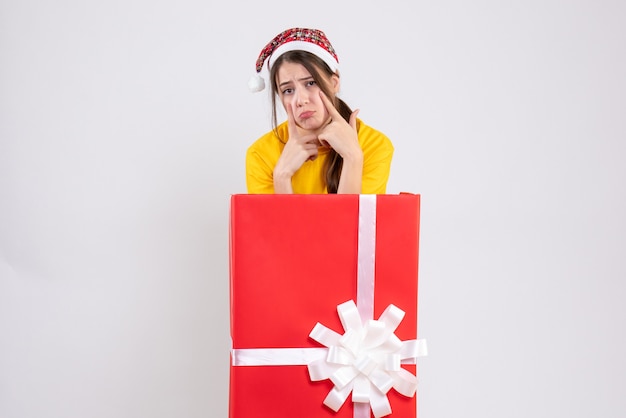 Niña triste con gorro de Papá Noel de pie detrás de gran regalo de Navidad en blanco