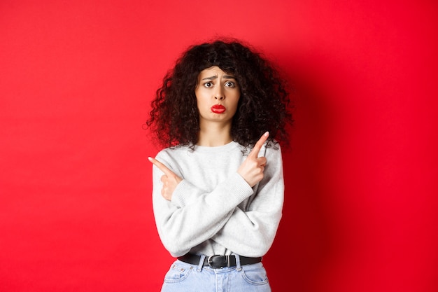 Niña triste nerviosa con el pelo rizado, sollozando miserablemente y señalando con el dedo hacia los lados, mostrando dos cosas y mirando indeciso, fondo rojo