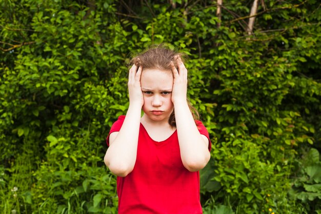 Niña triste que tiene dolor de cabeza mirando a la cámara de pie en el parque