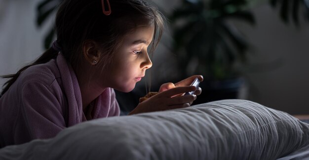 Niña usa un teléfono inteligente acostado sobre la almohada en casa