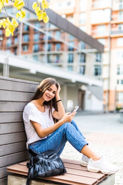 Niña usando un teléfono inteligente y mensajes de texto sentado en un banco de un parque urbano