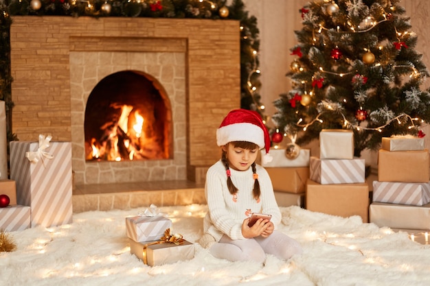 Niña usando un teléfono inteligente, revisando las redes sociales o jugando videojuegos, vestida con suéter blanco y sombrero de santa claus, posando en la sala festiva con chimenea y árbol de Navidad.