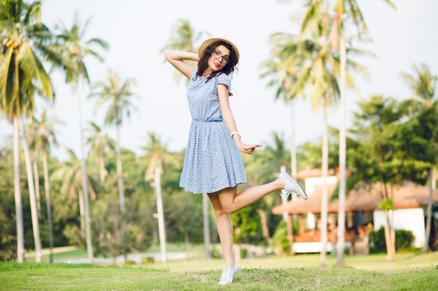 Niña con vestido azul cielo está de pie sobre una pierna en puntas de los pies en un parque. La niña tiene sombrero de paja y gafas negras.