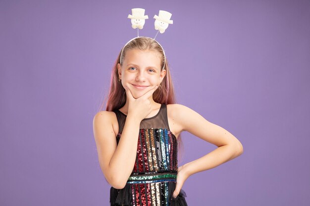 Niña en vestido de fiesta brillante y diadema divertida mirando a cámara con cara feliz sonriendo alegremente de pie sobre fondo púrpura