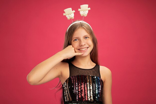 Niña con vestido de fiesta brillante y diadema divertida mirando a la cámara sonriendo haciendo gesto de llamarme, concepto de vacaciones de celebración de año nuevo sobre fondo rosa