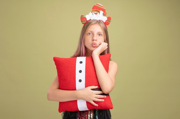 Niña en vestido de fiesta brillante y diadema con santa sosteniendo una almohada divertida con la mano en la barbilla esperando de pie sobre la pared verde