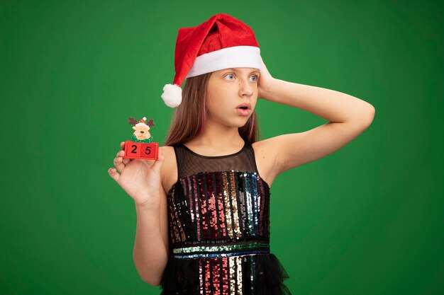 Niña en vestido de fiesta brillante y gorro de Papá Noel mostrando cubos de juguete con fecha veinticinco mirando a un lado sorprendido de pie sobre fondo verde