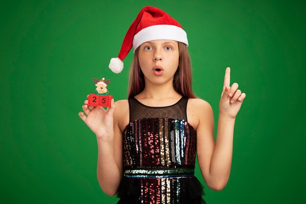 Niña en vestido de fiesta brillante y gorro de Papá Noel mostrando cubos de juguete con fecha veinticinco mirando sorprendido mostrando el dedo índice sobre fondo verde