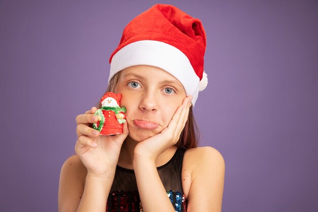 Niña con vestido de fiesta brillante y gorro de Papá Noel que muestra el juguete de Navidad mirando a la cámara haciendo una boca torcida con expresión decepcionada de pie sobre fondo púrpura