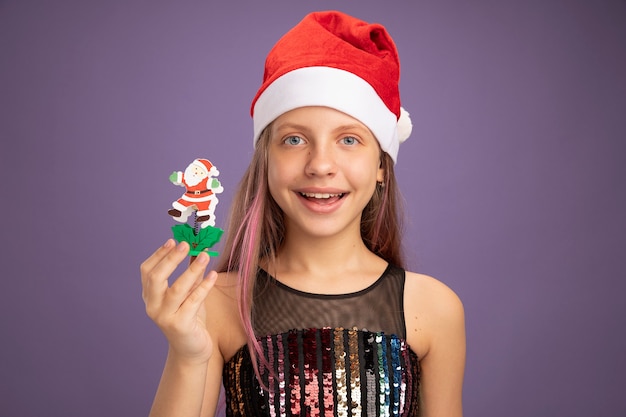 Niña en vestido de fiesta brillante y gorro de Papá Noel que muestra el juguete de Navidad mirando a la cámara con una sonrisa en la cara de pie sobre fondo púrpura