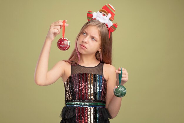 Niña en vestido de fiesta de brillo y diadema con santa sosteniendo bolas de navidad mirándolos confundidos teniendo dudas sobre fondo verde