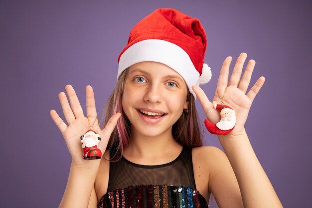 Niña en vestido de fiesta de brillo y gorro de Papá Noel con juguetes de Navidad mirando a la cámara con cara feliz sonriendo alegremente de pie sobre fondo púrpura