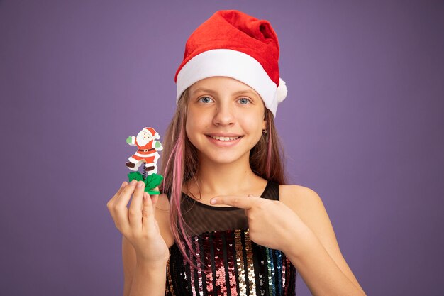 Niña en vestido de fiesta de brillo y gorro de Papá Noel que muestra el juguete de Navidad apuntando con el dedo índice sonriendo de pie sobre fondo púrpura