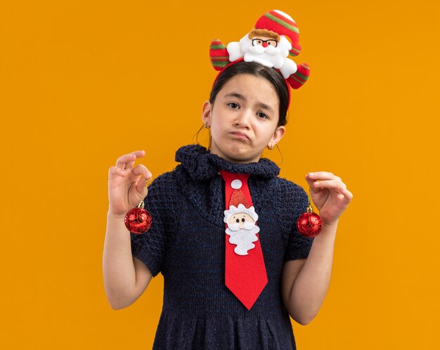 Niña en vestido de punto con corbata roja con borde gracioso en la cabeza sosteniendo bolas de navidad mirando confundido con expresión triste