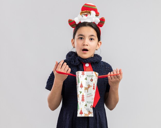 Niña en vestido de punto con corbata roja con divertido borde navideño en la cabeza abriendo una bolsa de papel con regalo de navidad mirando sorprendido