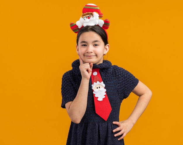 Foto gratuita niña en vestido de punto con corbata roja con divertido borde navideño en la cabeza mirando con expresión escéptica