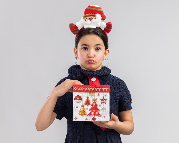 Niña en vestido de punto con corbata roja con divertido borde navideño en la cabeza sosteniendo un regalo de navidad mirando confundido