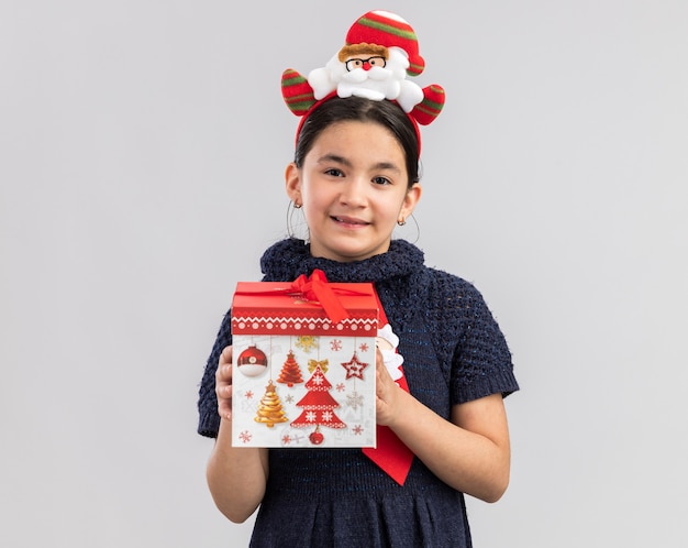 Niña en vestido de punto con corbata roja con divertido borde navideño en la cabeza sosteniendo un regalo de navidad mirando con una sonrisa en la cara feliz y positiva