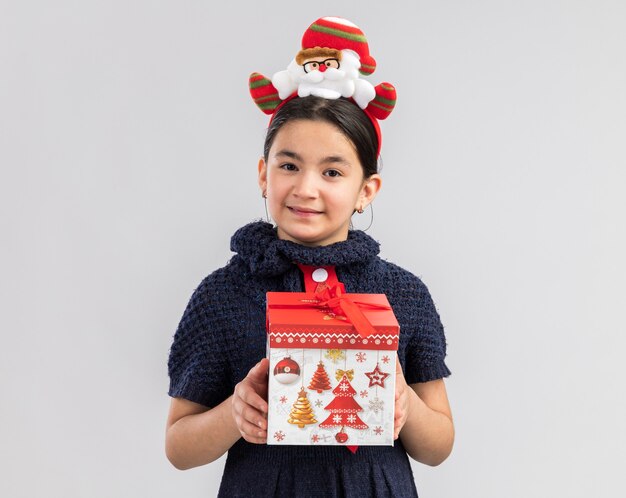 Niña en vestido de punto con corbata roja con divertido borde navideño en la cabeza sosteniendo un regalo de navidad mirando con una sonrisa en la cara feliz y positiva