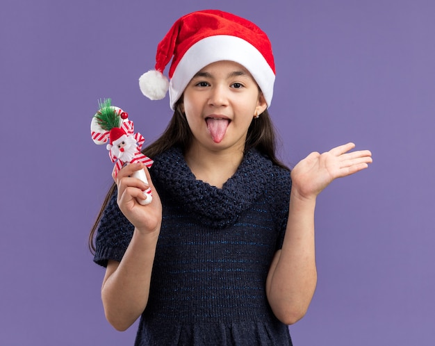 Niña en vestido de punto con gorro de Papá Noel con bastón de caramelo de navidad feliz y alegre sacando la lengua de pie sobre la pared púrpura