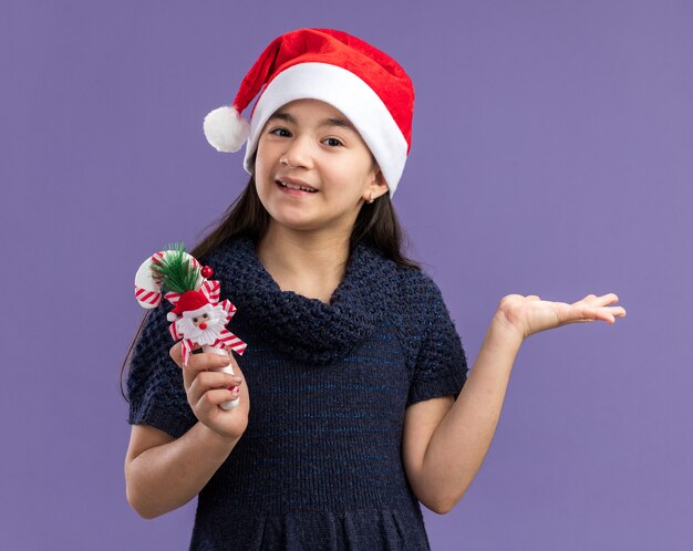 Niña en vestido de punto con gorro de Papá Noel con bastón de caramelo de Navidad con una sonrisa en la cara feliz y alegre de pie sobre la pared púrpura