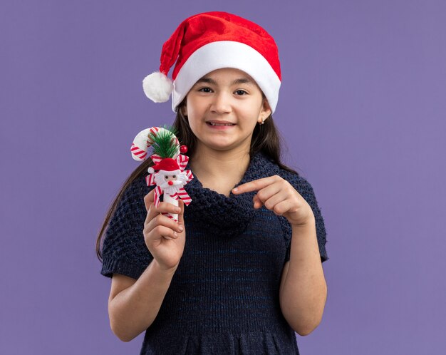 Niña en vestido de punto con gorro de Papá Noel sosteniendo bastón de caramelo de Navidad apuntando con el dedo índice a él feliz y positivo sonriendo alegremente de pie sobre la pared púrpura