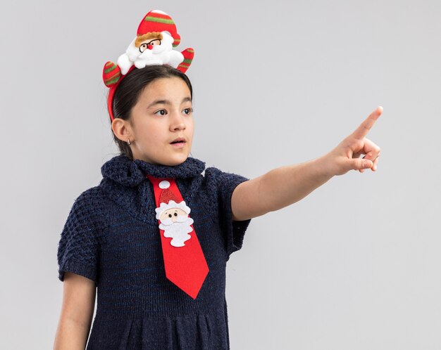 Niña en vestido tejido con corbata roja con gracioso borde navideño en la cabeza apuntando con el dedo índice a algo a un lado preocupado