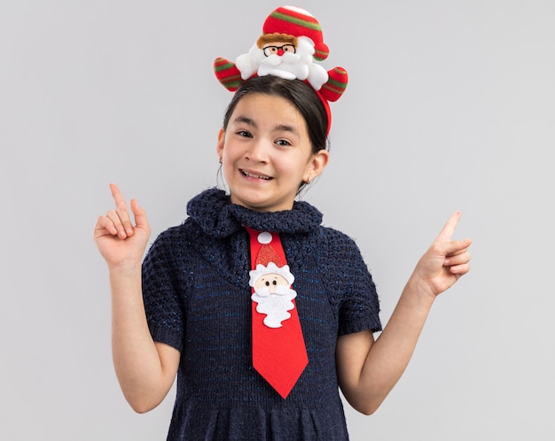 Niña en vestido tejido con corbata roja con gracioso borde navideño en la cabeza, feliz y positivo sonriendo mostrando los dedos índices