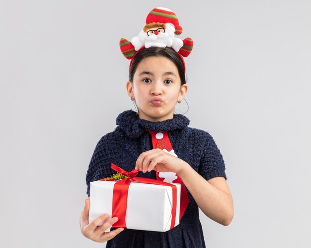 Niña en vestido tejido con corbata roja con gracioso borde navideño en la cabeza sosteniendo el regalo de Navidad mirando confundido