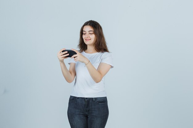 Niña viendo videos en el teléfono móvil en camiseta, jeans y mirando feliz, vista frontal.