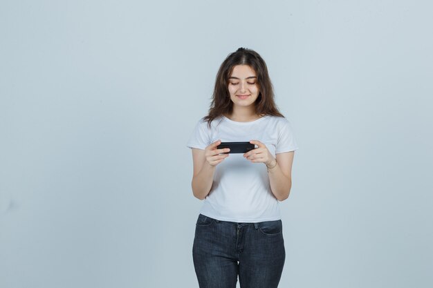 Niña viendo videos en el teléfono móvil en camiseta, jeans y mirando feliz, vista frontal.