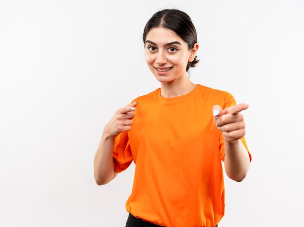 Niña vistiendo camiseta naranja apuntando con los dedos índices sonriendo alegremente con cara feliz de pie sobre la pared blanca