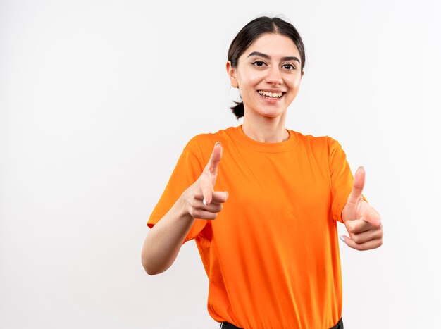 Niña vistiendo camiseta naranja apuntando con los dedos índices sonriendo alegremente de pie sobre la pared blanca