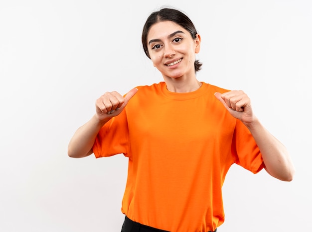 Niña vistiendo camiseta naranja apuntando a sí misma sonriendo confiada feliz y positiva de pie sobre la pared blanca